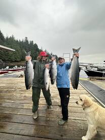 Two young anglers proudly displaying their catch of large salmon at the dock, surrounded by a scenic backdrop of trees and boats.