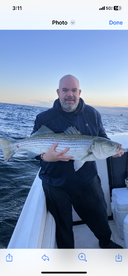 Angler proudly displaying a large Striped Bass while fishing on a boat during sunset.