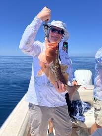Angler proudly displaying a large Grouper caught during a deep-sea fishing trip, with a clear blue sky and calm waters in the background.