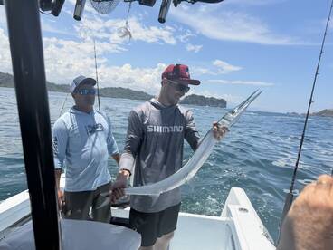 Angler proudly displaying a large Barracuda while fishing on a boat in a scenic coastal environment.