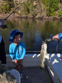 A young angler smiles while holding a freshly caught Speckled Trout on a fishing trip, showcasing the excitement of fishing in a scenic river environment.