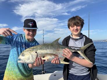 Two young anglers proudly displaying their catch, a large fish, while fishing on a boat in clear blue waters.