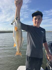 A young angler proudly displaying a Redfish caught during a fishing trip, with a scenic backdrop of water and sky.