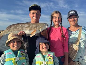 A family proudly displays a large Redfish caught during their fishing trip, showcasing smiles and excitement on a sunny day.