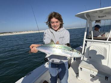 A young angler proudly displaying a vibrant Spanish Mackerel while fishing from a boat in clear waters.
