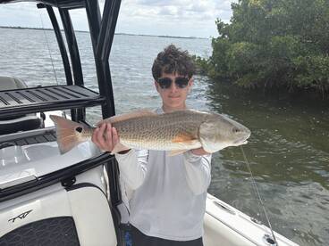 A young angler proudly displaying a large Redfish while fishing from a boat in a scenic coastal environment.
