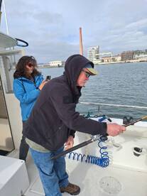 Two anglers on a boat, one actively reeling in a catch while the other captures the moment with a camera, showcasing a lively fishing experience.