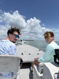 Two anglers enjoying a sunny day on a fishing boat, with a clear view of the water and clouds in the background.