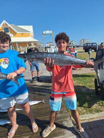 Two young anglers proudly displaying their catch at the marina, showcasing a large King Mackerel after a successful fishing trip.