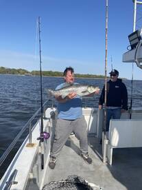 Angler proudly displaying a large Striped Bass while fishing on a charter boat, with another fisherman in the background.