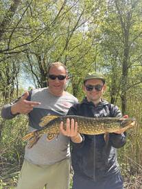 Two anglers proudly displaying a Northern Pike caught during a fishing trip in a lush, green environment.