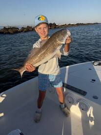 Young angler proudly holding a large Redfish while standing on a boat, showcasing a successful fishing trip.