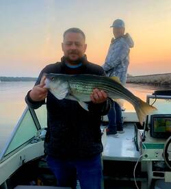 Angler proudly displaying a large Striped Bass while fishing at sunrise on a tranquil lake.