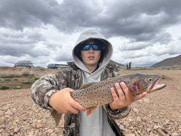 A young angler proudly displaying a large trout against a backdrop of cloudy skies and rocky terrain.