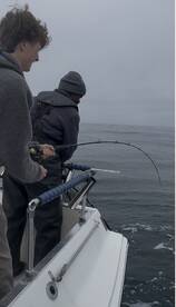 Two anglers engaged in reeling in a fish while fishing on a boat in overcast conditions.