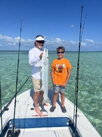 A young angler and an adult proudly displaying a large Barracuda on a boat in clear waters, showcasing a successful fishing trip.