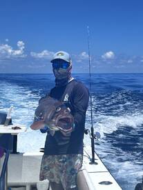 Angler proudly displaying a large Grouper while fishing offshore, with a clear blue sky and ocean in the background.