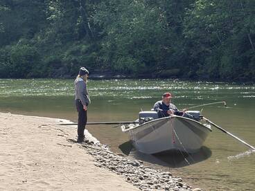A man fishing from a small boat while a woman stands on the shore, enjoying a peaceful day on the river surrounded by lush greenery.