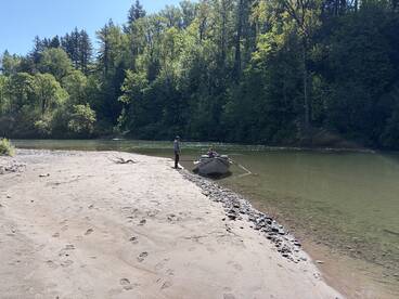 Two anglers enjoying a peaceful day on a river, with a drift boat anchored on the sandy shore surrounded by lush greenery.