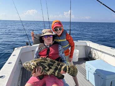 Two young anglers proudly displaying a large Grouper while fishing on a boat in open waters.