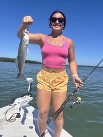 A young woman proudly displaying a Speckled Trout while fishing from a boat in a sunny coastal environment.