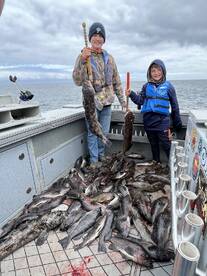 Two young anglers proudly displaying their catch of multiple species, including large cod, on a fishing boat in a coastal environment.