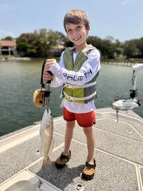 A young angler proudly holding a freshly caught Redfish while standing on the deck of a boat in a scenic fishing environment.