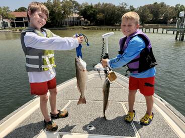 Two young anglers proudly displaying their catches, a Redfish and a Trout, while enjoying a day of fishing on a calm waterway.