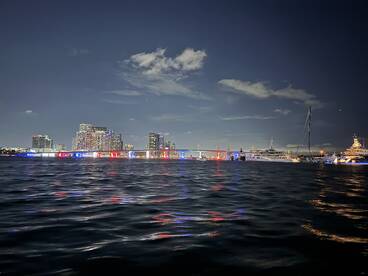 Nighttime view of a city skyline with illuminated buildings and boats on the water, creating a vibrant atmosphere.