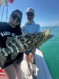 A smiling angler proudly displays a large Goliath Grouper while fishing in clear waters, showcasing a successful day on the boat.