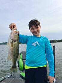 A young angler proudly displaying a large Largemouth Bass while fishing from a boat on a serene lake.