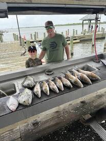 A father and son proudly stand by a cleaning station displaying a variety of fish, including Redfish and Flounder, after a successful day of fishing at the dock.