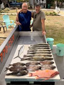 Two anglers proudly displaying their catch of various fish, including a large Red Snapper and several Trout, arranged on a cleaning table after a successful fishing trip.