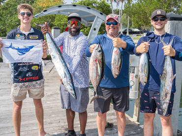 A group of anglers proudly displaying their catch, including a large Kingfish and several smaller fish, at the dock after a successful fishing trip.