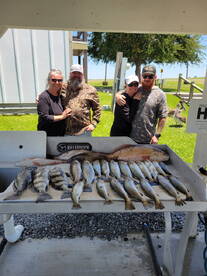 A group of four anglers proudly posing with their catch of various fish, including Speckled Trout, displayed on a cleaning table after a successful fishing trip.