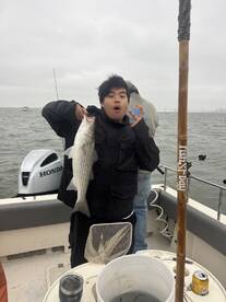 A young angler excitedly holds up a striped bass while fishing on a boat, showcasing a successful catch during a cloudy day.