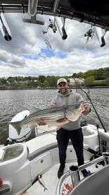 Angler proudly displaying a large striped bass while fishing from a boat on a calm river.