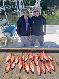 A couple proudly stands beside a catch of redfish displayed on a wooden board after a successful fishing trip.