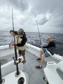 Two anglers actively fishing on a charter boat, with one preparing to reel in a catch while the other waits with a rod in hand, set against a cloudy ocean backdrop.