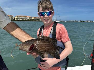 A young angler proudly displaying a caught fish while fishing on a sunny day, with a scenic waterfront in the background.