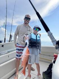 A young angler proudly displays a large Red Snapper while fishing with a family member on a boat.