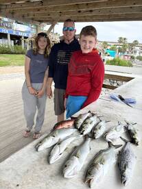 A family proudly displays their catch of various fish, including Snapper and Trout, at a dockside cleaning station after a successful fishing trip.