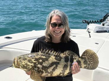 A smiling angler proudly displaying a large Grouper while on a fishing boat, showcasing a successful day on the water.