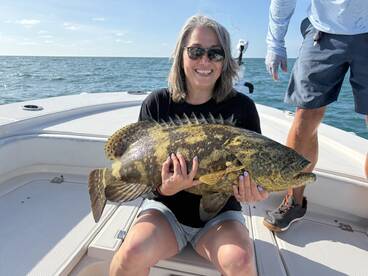 A smiling angler proudly displays a large Goliath Grouper while fishing on a boat in clear waters.