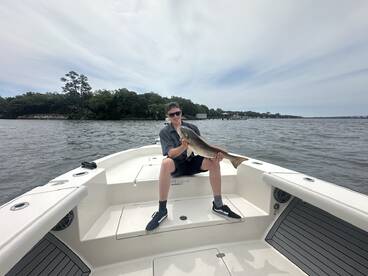 A young angler proudly displaying a large Redfish while seated on a boat in a scenic waterway.