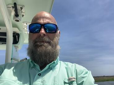 A bearded angler takes a selfie inside the boat, showcasing the helm area and clear blue skies.