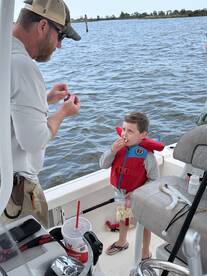 A young boy in a life jacket enjoys a snack while an adult prepares bait on a boat, showcasing a family-friendly fishing experience.