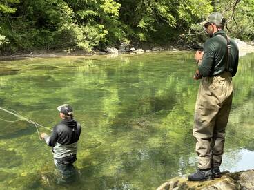 Two anglers fishing in a serene river setting, one casting while the other observes, showcasing the tranquility of fly fishing.