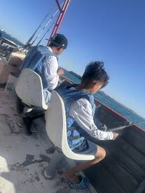 Two young anglers seated on a boat, focused on fishing in a sunny coastal environment.