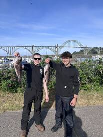 Two young anglers proudly displaying their catch of rockfish in front of a scenic marina and bridge backdrop.